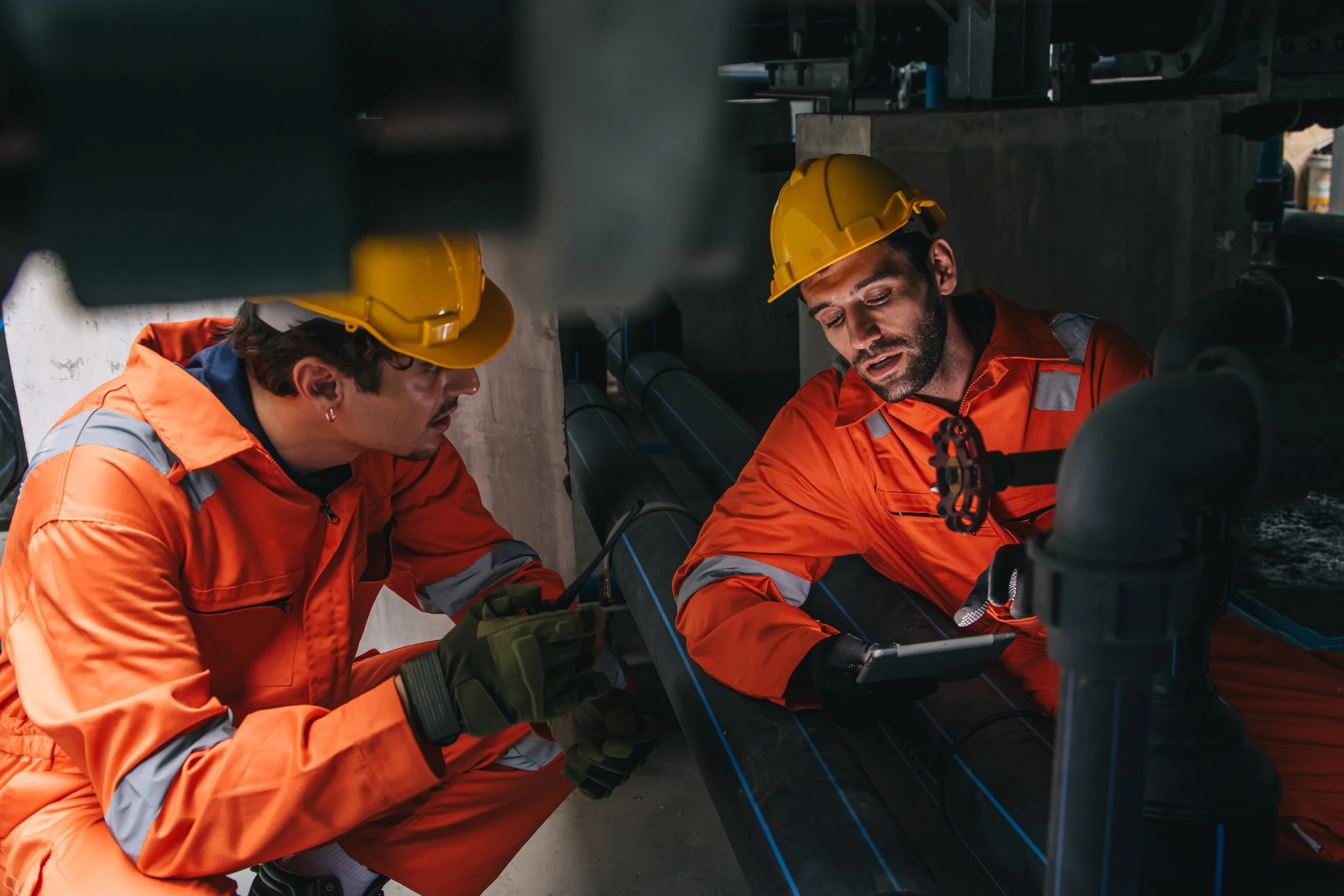 Two workers in orange high-visibility overalls and hard hats crouched over industrial pipework, one holding a tablet