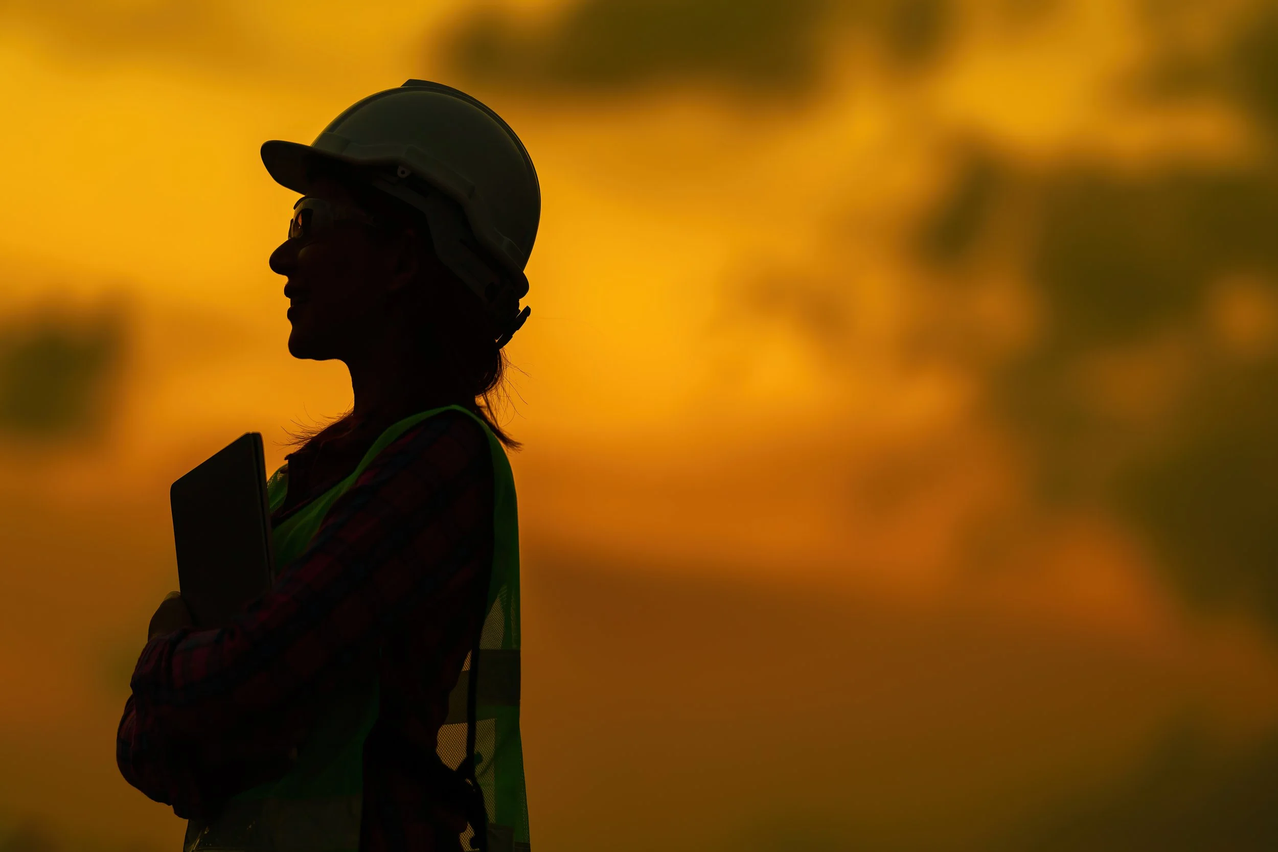 A worker in a hard hat and high-visibility vest holding a tablet, silhouetted against an orange sunset on site